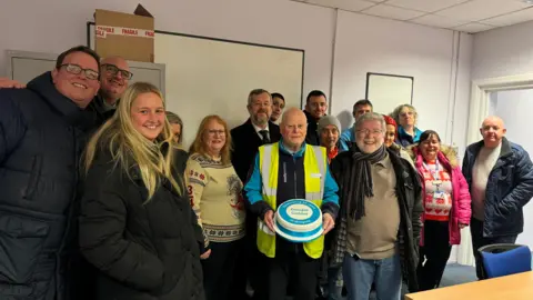 A group of around 15 people all smiling at the camera. In the centre of the image is Mr Calder. He is wearing a bright yellow Hi-Viz jacket and is holding a cake. 