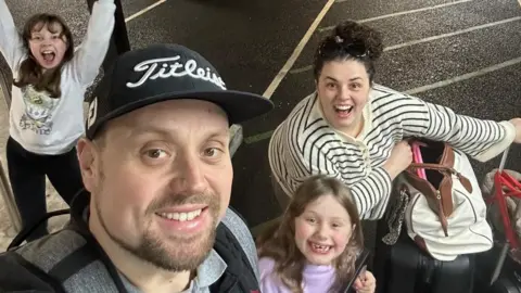 Family Harry, Brydee, Eliza and Lillie are in a car park with their luggage for their flight out from Heathrow. They are all looking up at the camera in the selfie and smiling with excited expressions.