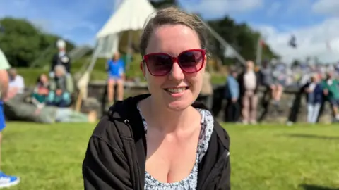 Johanna Downing-Green has her hair tied back and is wearing sunglasses, sitting in front of Tynwald Hill, smiling on a sunny day.