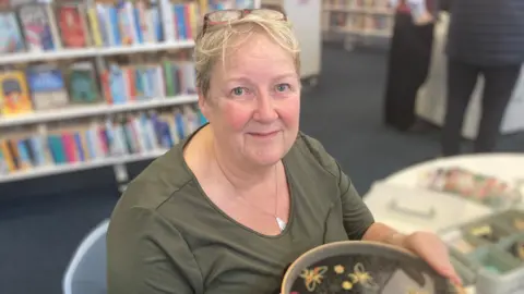 Laura Coffey/BBC A woman with a dark green top and silver necklace is sitting at a table. She has a pair of glasses propped on her head. She has blonde hair. Behind her are shelves of books.