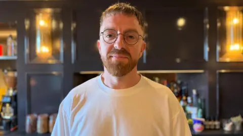 An image of Gareth Evans standing in the hotel bar area - he has a brown beard, glasses and is wearing a white T shirt