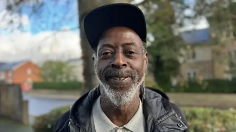 Dexter Stewart, better known as Papa D, smiling at the camera. He is wearing a cap, black coat and grey polo shirt.