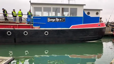 Jon Wright/BBC A side view of a barge, with a blue painted woodwork, with red edging and a black hull. A sign on the side reads Barge No. 9. A group of people stand in high-visibility jackets on the deck. 