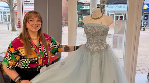 A woman smiles as she holds a princess dress, which is baby blue with glittery embellishments, on a mannequin in a shop window, with a high street outside. The woman wears large glasses and a bold-patterned multi-coloured shirt. She has prominent facial piercings. 