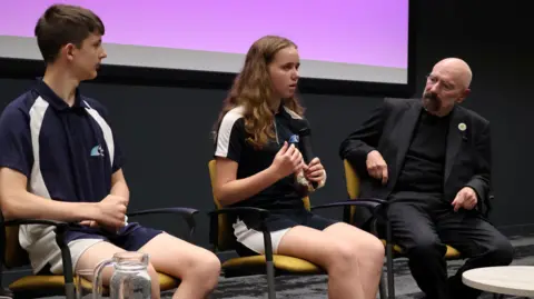 Jon Wright/BBC Two high school students in sports uniform sit on chairs at the front of a hall, with a man in suit looking at them.