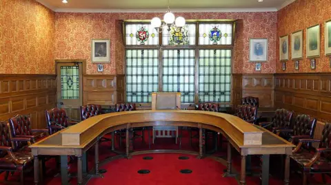 MANX SCENES The Legislative Council chamber, which has wooden panelled walls and features a horseshoe shaped wooden table with chunky leather clad chairs. There are portraits of former governors on the wall on the right and large windows with stained glass emblems at the top.