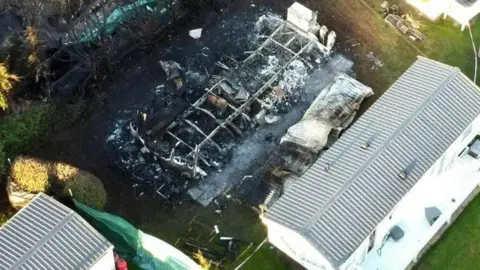 An aerial view of the remains of a caravan that has been on fire. You can see its foundations but there is a lot of black soot and nothing left over. Other caravans can be seen next to it.
