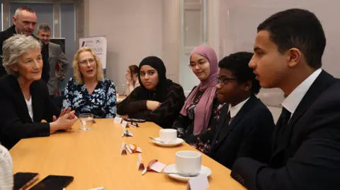 PA Media Shows a women in grey hair and black jacket on the left, next to a woman in blond hair and blue, black and white shop and four young people - two girls and two boys. The girls are wearing headscarves. 