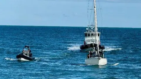 National Crime Agency The picture is taken out on the open sea with blue sky behind. Three boats are heading straight towards the camera. The yacht has it's sails down and is being escorted into Haslar Marina by a rigid inflatable boat on the left of the picture and a bigger border force vessel behind it. 