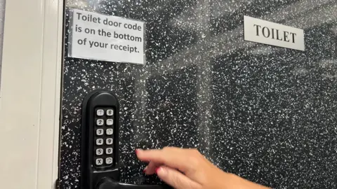 A hand pointing to a number pad on the lock of a black pebbledash door, which has a sign reading "Toilet" and another reading "Toilet door code is on the bottom of your receipt".