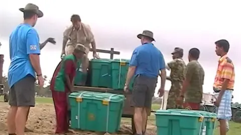 Shelterbox A group of men pull large green plastic boxes off a cart.