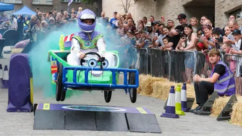 A man is driving a small kart, which is flying in the air over a ramp on a grey brick path. There are spectators watching behind metal barriers. The kart it green and blue and is decorated with two eyes on the front. The man is wearing a Buzz Lightyear, purple green and white race suit with a purple helmet. There is blue smoke coming from the back of it
