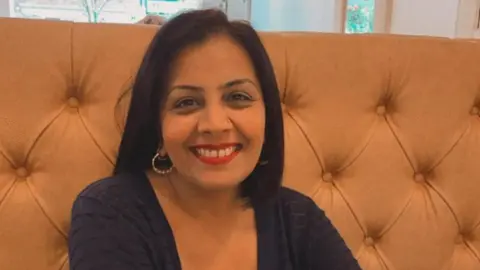 A woman with dark hair and wearing a dark top is sitting down and smiling at the camera. Part of a window is at the top of the photo.