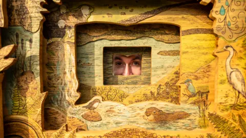 West Cumbria Rivers Trust Artist Daniel Weatheritt is peeking through the sculpture, which has an aperture-like opening. It has sheep, beavers and birds carved on it.