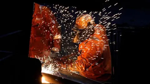 Someone in a hi-vis orange jacket and helmet welding. There are sparks flying from some steel in front of them. There is another person in a hi-vis orange jacket to the left.