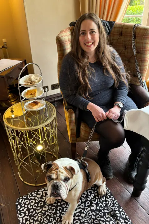 Vanessa Ward Photograph of Vanessa Ward, 37, pictured sat on an armchair in a restaurant. There is a high tea on a table next to her, and her bulldog, bear, sits on a rug infront of her, by her feet. She has brown long hair down to her waist and wears a grey long sleeve dress and black sheer tights. 