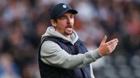 PA Media Joey Barton, who is wearing a grey hoodie and gillet, gesticulates from the touchline at a football match. 