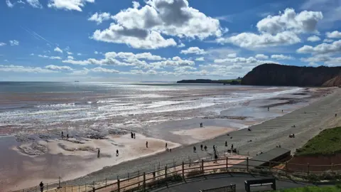 BBC Weather Watchers/Tony N-D The beach at Sidmouth on a bright day. There are about 20 people on the beach, some with dogs. The clouds are reflected in the water and the waves are quite small. The beach is sandy near the waterline and stony beyond it. 