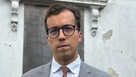 A man with dark hair and dark rimmed circular glasses wearing a grey jacket, white shirt, and checked yellow and navy blue tie, standing in front of a white wall and boarded up window.