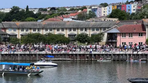 Hundreds of people are sat along Bristol Harbour, with boats on the water and coloured houses and trees in the background.