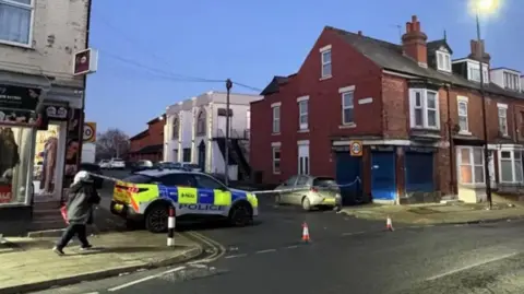 A road is closed off with cones in the road and police tape across the top of the street. There is a marked police car parked across the top of the street 