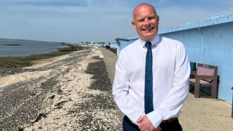 James Mason in a shirt and tie standing on Canvey beach with dark stones on a slant past his right shoulder and a blue wall and benches to his left.