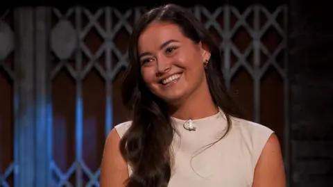 Hair Syrup A young woman with long brown hair and a beige top smiling.