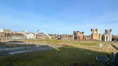 BBC A view of the grassy field where the arena is supposed to be built. The white, curved Millennium bridge is poking out from over the hill along with the red and yellow Baltic art gallery.
