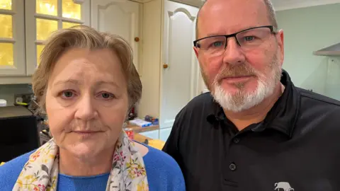 Nadine and Rob Thomas look at the camera standing in their kitchen. Nadine wears a charcoal blue jumper with a while scarf decorated with flowers, and Rob wears a black t-shirt.