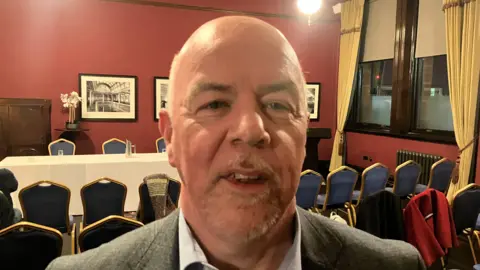 A bald man with a white goatee beard pictured in a red walled committee room with a large table and numerous blue chairs around it. There is a window with large gold curtains.