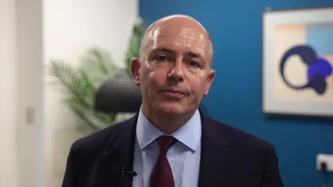 Roland Sinker wearing a dark suit jacket with a white shirt and a deep red tie. He is indoors in front of a blue wall. Behind him, there is a large framed abstract artwork. A green plant and a black floor lamp are visible behind him. His head is tilted slightly as he looks directly at the camera.