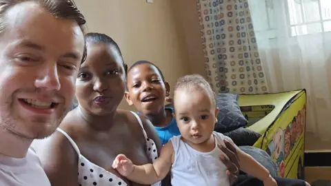 Submitted A family of four smiles for a photo inside a living room. A man, a women, and their two young children aged 11 and two. They seem happy together.