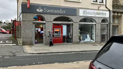 Looking across the street to the Santander branch. An elderly woman is walking past. 