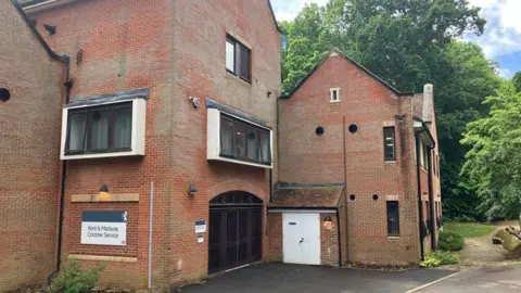 Exterior of a red-brick coroner’s service building with multiple windows, a driveway entrance and a sign reading “Kent & Medway Coroner Service”, with trees behind.