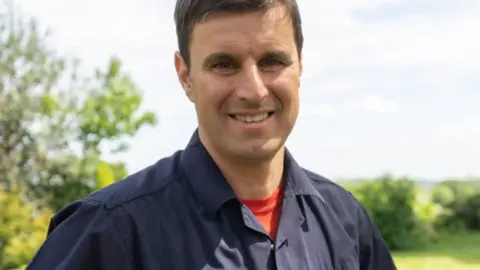 Glynn Luznyj, a man wearing a red t-shirt and navy blue firefighter overalls. He is standing outdoors with fields and trees behind him.