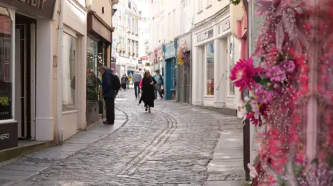 Guernsey's High Street. A cobbled street with a few people walking on it and people either side. 
