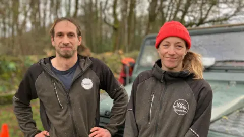 A man and a woman - both tree surgeons - are standing in front of a 4 by 4 in a woodland. They are wearing work wear which is covered in bits of sawdust. One wears a bright orange hat. The circular branding on their fleeces says the company name TreeWise