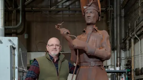 University of Sunderland Artist Ron Lawson next to Molly the statue. He is wearing a check shirt and green gilet. Molly is about two foot taller than him. She is brown and is holding a welding torch.