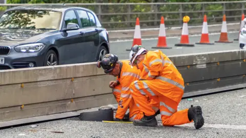 Two men working on a repair on the motorway. They are wearing orange high-vis tops and bottoms, each donning a hard hat. One is reaching into a hole they have created in the concrete. On the other side of the carriageway, across the concrete barrier, cars continue to flow through - but a lane has been cordoned off with orange cones.