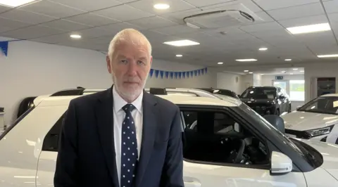 Martin Gurren is pictured standing in a car showroom. A white car is visible behind him. He's wearing a blue suit with a blue polka dot tie. He has white hair and a white beard.