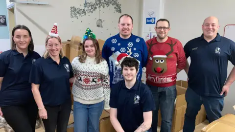 Three women and four men, some wearing Christmas jumpers and hats, others wearing blue polo shirts, stand in a line in front of a stack of brown boxes in a hospital lobby.