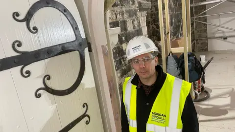 Shaun Whitmore/BBC James Spedding is wearing a high-vis jacket and a white helmet stood next to a large wooden door with steel braces and decorations