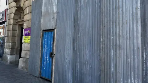 Alex Pope/BBC Corrugated metal hoarding lines the pavement outside a library building, with a small blue door set into the barrier.
Temporary metal panels cover part of a stone library frontage, with a sign visible beside an entrance.
A narrow street view showing construction hoarding attached to a building, with a blue access door and signage for library opening hours.
Metal sheeting encloses building works on a town-centre library, partially obscuring the entrance.
A pavement bordered by grey construction panels, with a small secured doorway and library signage behind.