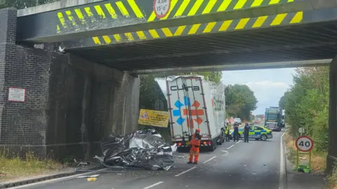 A road under a railway bridge. There is debris under the bridge and two emergency service workers. To the left there is a lorry with its roof damaged.