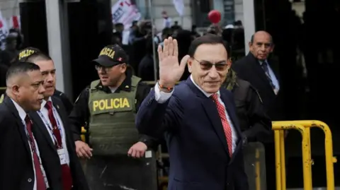 Reuters Martín Vizcarra in suit and tie and dark sunglasses smiles and waves as he walks past police on his way to court