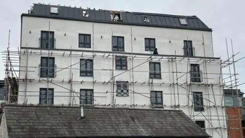Suki Somal/BBC A six storey building with white or cream rendering and scaffolding around it. It is cloudy above.
