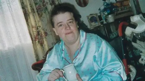 Family handout Angela Lloyd holds a white jug and smiles at the camera. She is sitting in her wheelchair and is wearing blue silk pyjamas and appears to be in her living room with floral curtains and various nick nacks on shelves behind her.