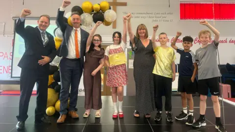 North Northamptonshire Council Eight people, all with their arms raised, looking happy and at the camera. Two men on the left are dressed in dark suits, with ties on, then five children are dressed in sports kits, one girl in a dress and one girl in a gold jumpsuit. A woman is wearing a black and white dress.