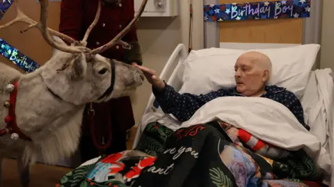 St Helena Hospice A man with no hair in a hospital bed strokes the nose of a reindeer.