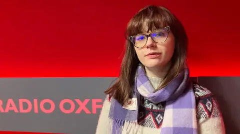 Megan poses for a picture in the radio studio. She has brown hair, cat-eye shaped glasses, and wears a purple checked scarf and grey patterned knitted jumper.